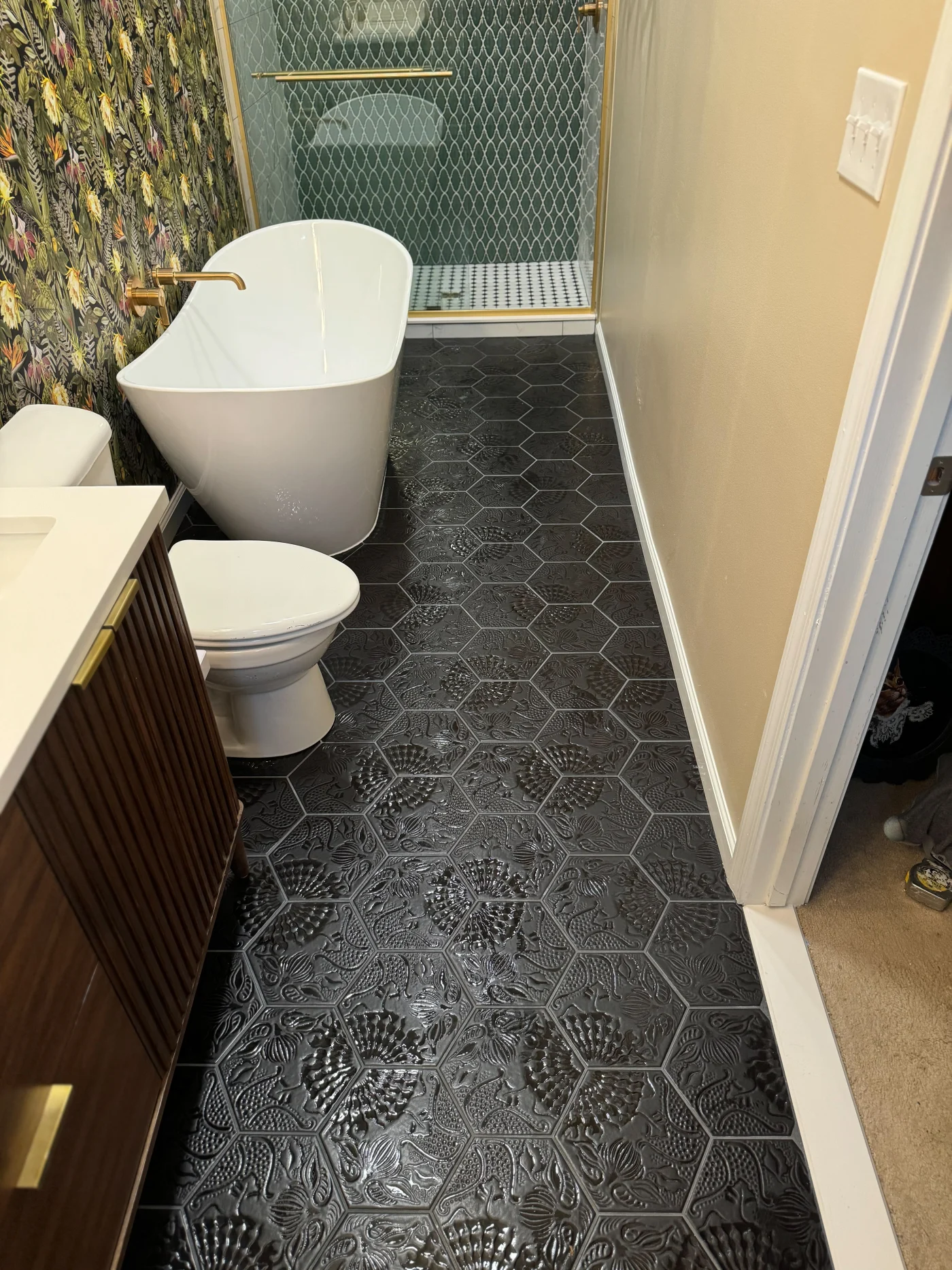 Spectacular Full Bathroom Remodel | Frankford, DE A remodeled bathroom featuring a freestanding white tub, patterned hexagon black tile flooring, gold fixtures, a floral accent wall, and a glass shower.