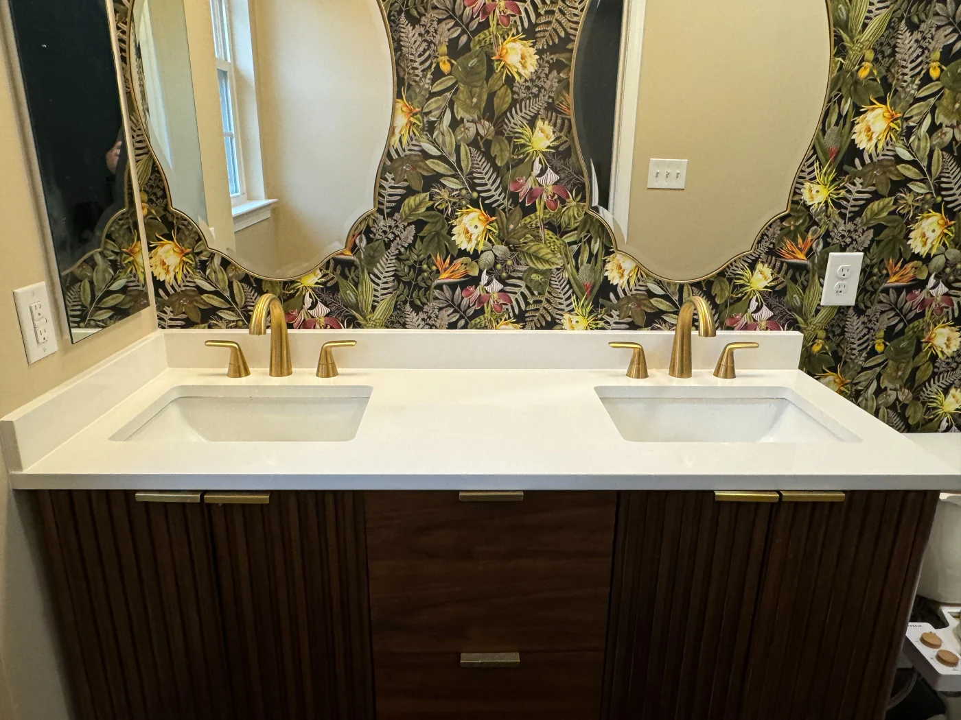 Quick 5 Day Bathroom Bethany Beach De Mast Contracting Double-sink bathroom vanity with a white countertop, brass faucets, and dark wood cabinetry, set against bold floral-patterned wallpaper.