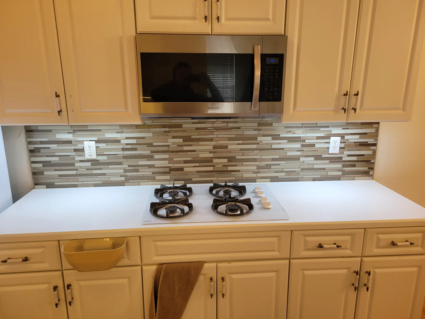 A white kitchen with glass tile backsplash, built-in microwave, gas cooktop, white countertops, and classic cabinet hardware.