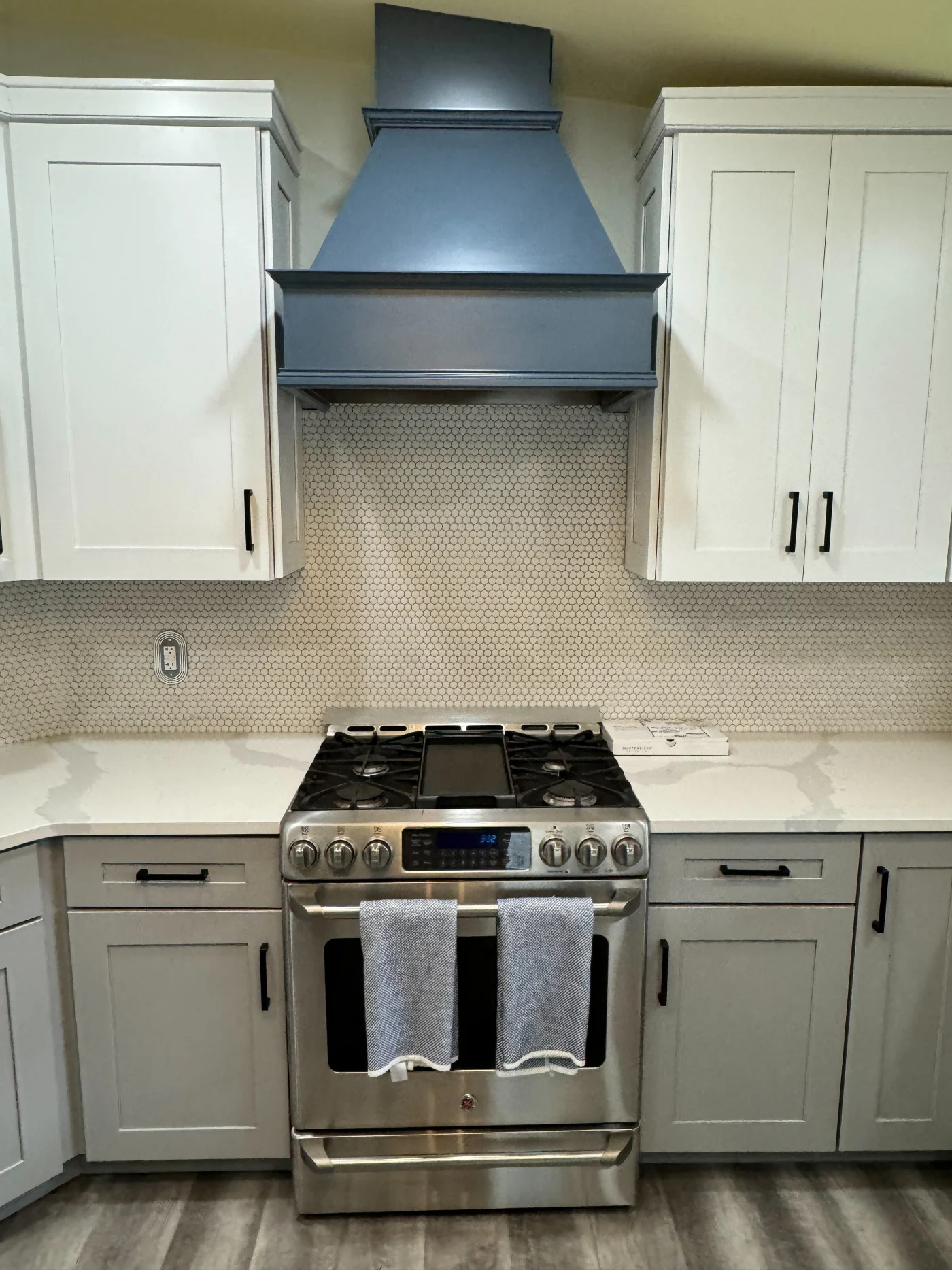 A modern kitchen with gray shaker cabinets, white countertops, stainless gas range, and a dark vent hood over a penny tile backsplash.