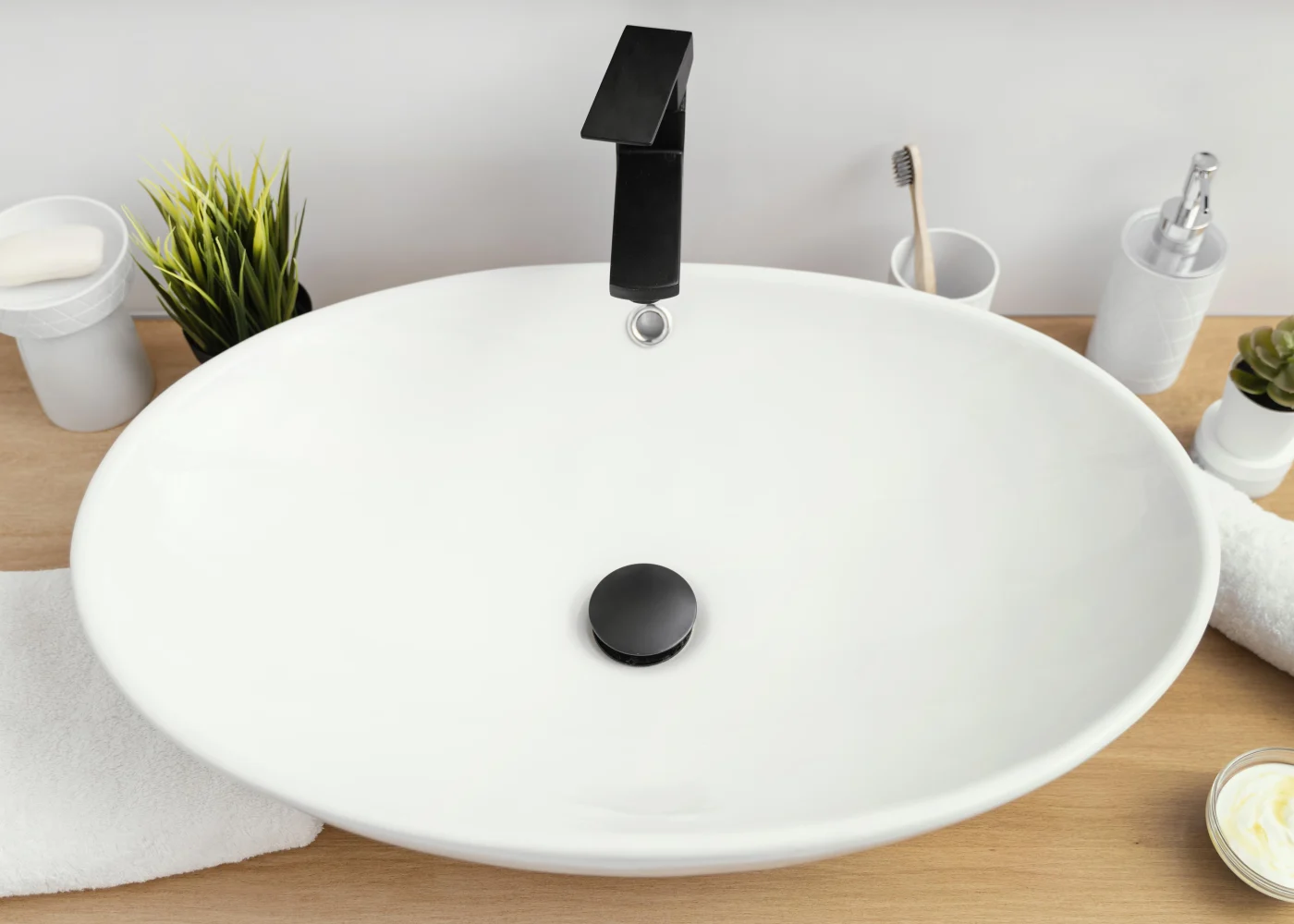 Gleaming 5-Day Bathroom Remodel Ocean View DE | Mast Contracting A white, oval shaped sink with a black faucet and stopper sits on a light brown wooden countertop. Surrounding the sink are bathroom accessories, including a toothbrush in a cup, a soap dish, a separate dispenser, small potted plants, and white towels.