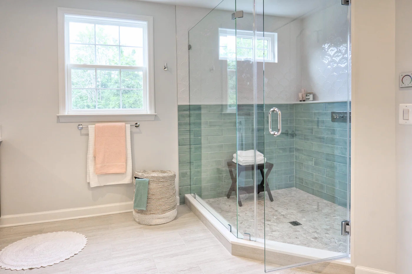 Smart Full Bathroom Remodel Ocean View, DE | Mast Contracting On the left, a large window takes up much of the light-gray colored wall, and there is a circular white rug on the floor. On the right, a glass-enclosed walk-in shower features two tone tile: white on the top half, and an ocean blue on the bottom.