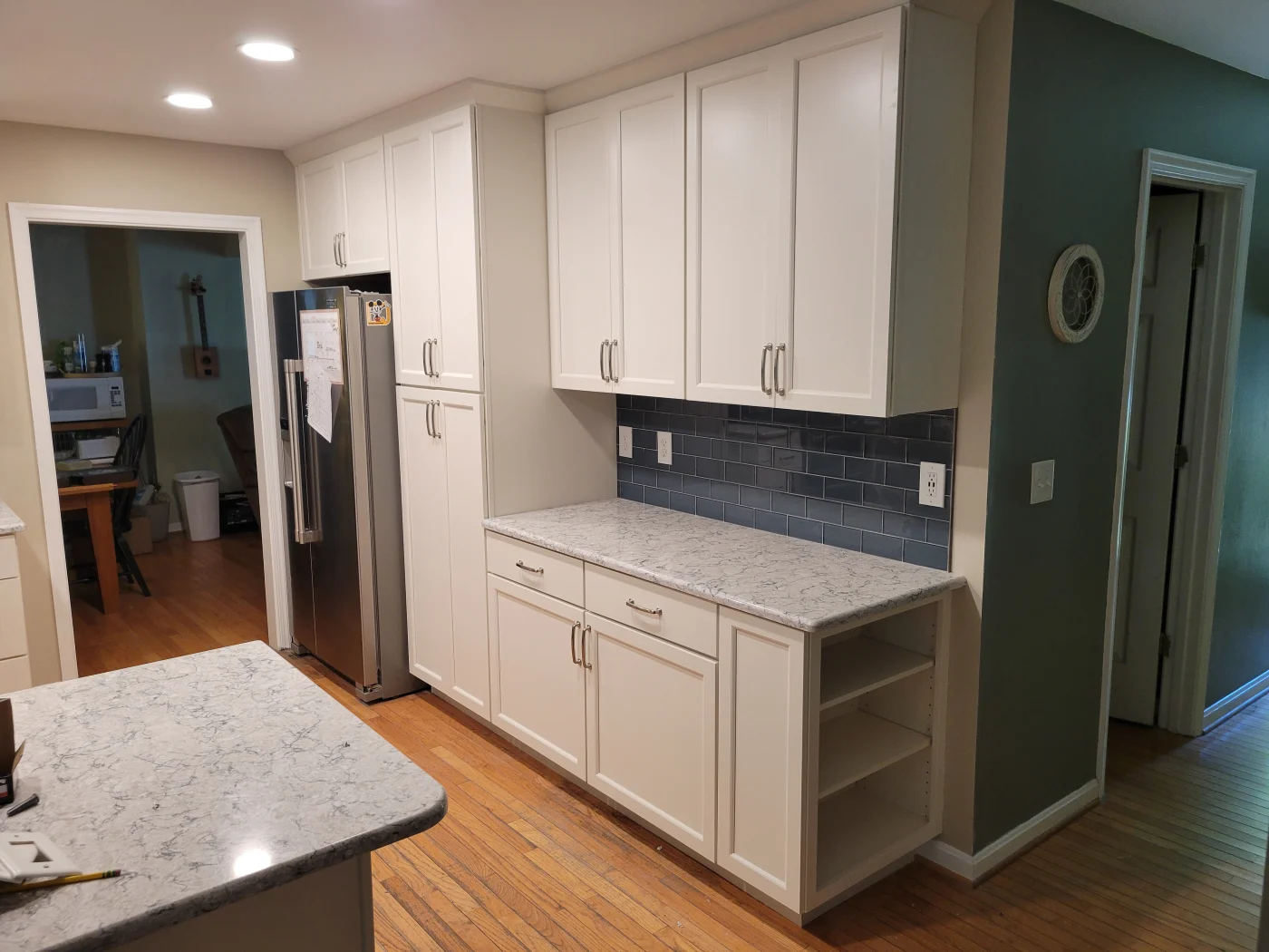 Deliberate Kitchen Remodeling Rehoboth Beach De Mast Contracting A photo of a home’s kitchen. There is a small island on the left, and the right wall is covered with upper and lower sets of cabinets. The cabinets are white, and the countertops are a gray and white marble pattern. There is blue subway tile on the wall between the cabinets, and the other walls are painted tan.