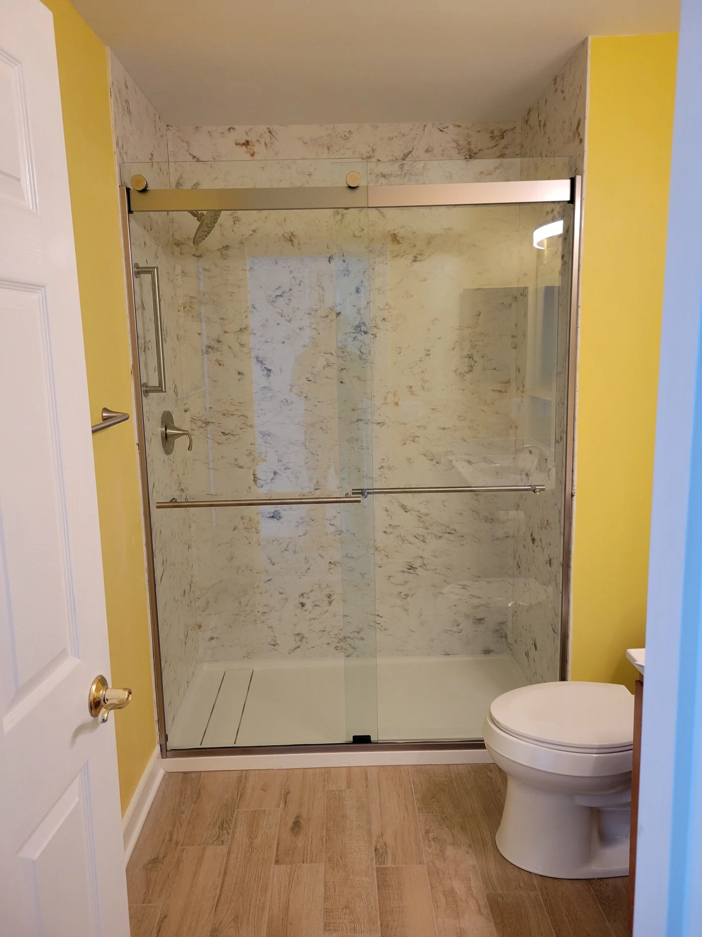 Bathroom And Kitchen Remodeler In Lewes, DE | Mast Contracting A photo looking into the bathroom and showing a glass-enclosed walk-in shower. The floor of the shower is white, and the walls are white with black splashes. The bathroom's walls are painted yellow, and the floor is light brown.