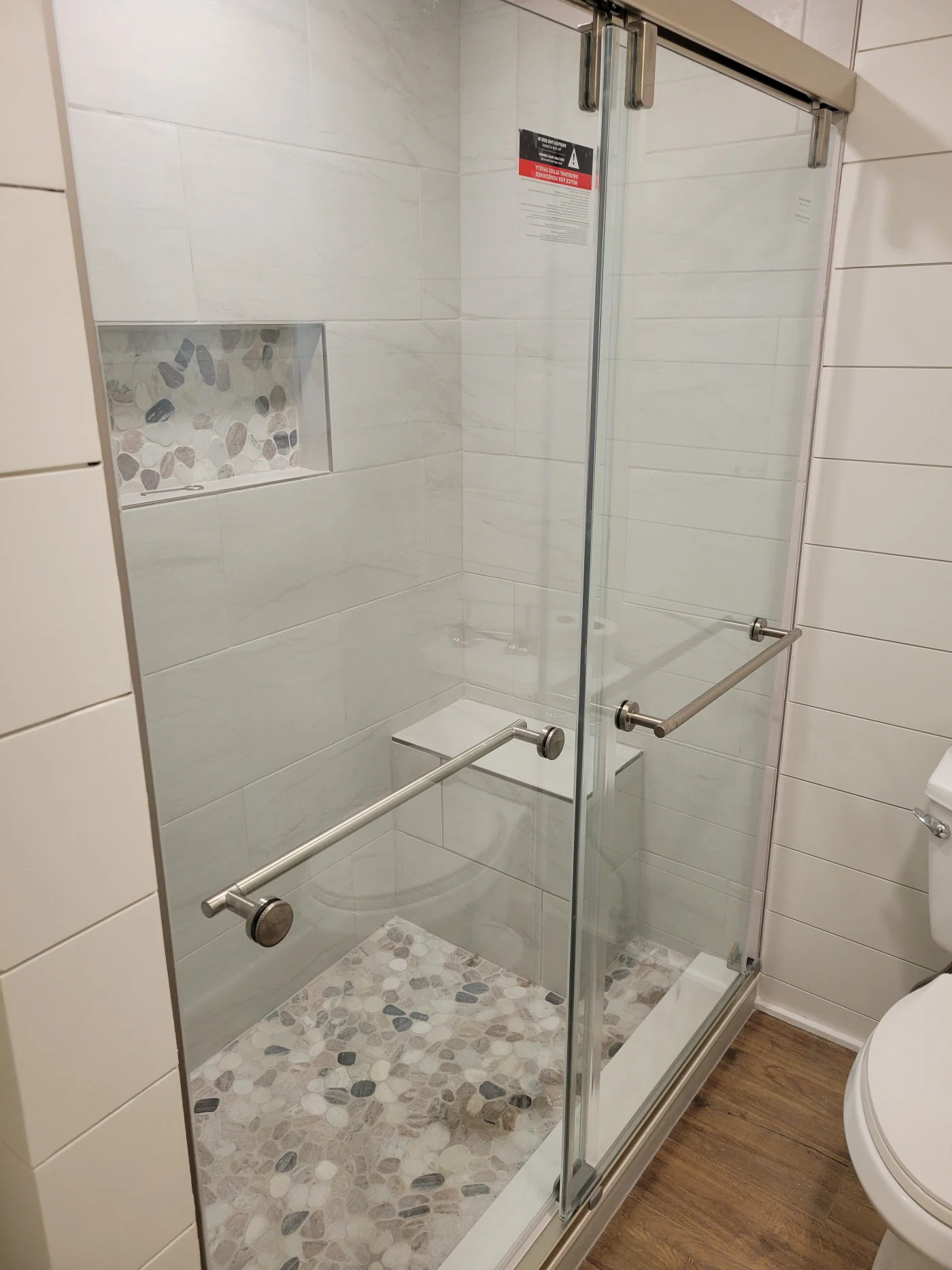 Honorable Bathroom And Kitchen Remodeler In Lewes, DE | Mast Contracting A step-in shower with large white tile on the walls, and a small bench seat on the right side. There is a cutout with natural stone tile along the back side, which matches the tile on the shower floor. The actual flooring of the bathroom is dark brown wood grain.