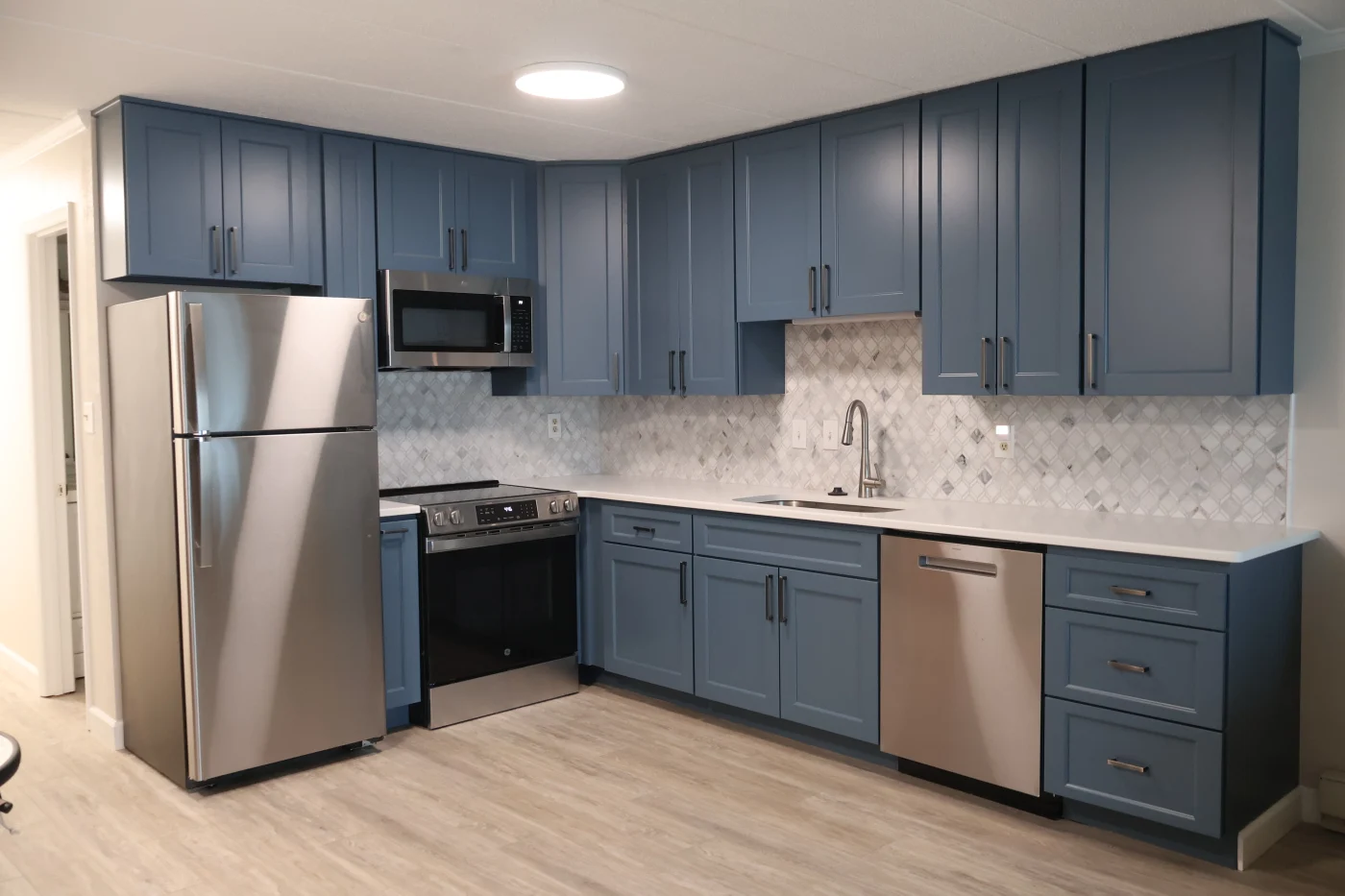 A large, open kitchen with dark blue cabinets, white countertops, and a light brown floor. There is a tile backsplash between the countertop and upper cabinets, and it's gray, white, and blue. The appliances are stainless steel, but the fridge and microwave have black accents.