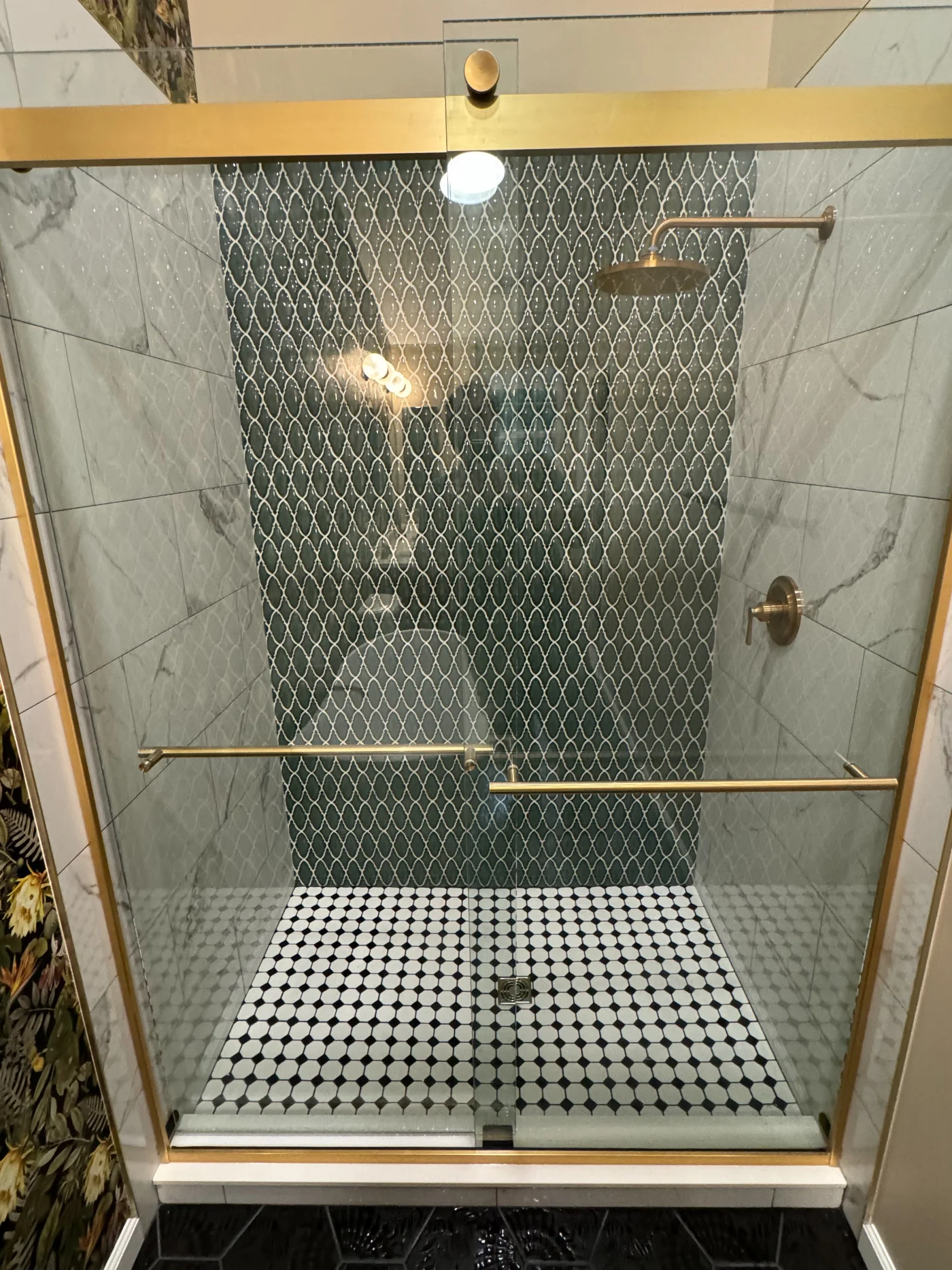 A square shower with a glass door, golden framing, and fixtures. The shower's floor is white circles with black grout, and the tile on the interior shower walls is a mixture of gray and white marbled and geometric black and white.