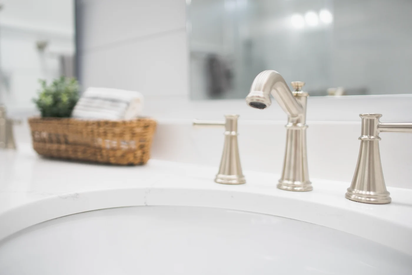 A close-up shot of a sink's silver faucet and handles. The sink and countertop are white, as is the wall behind the mirror mounted above the sink. There's a wicker basket with a small green plant to the left of the sink.