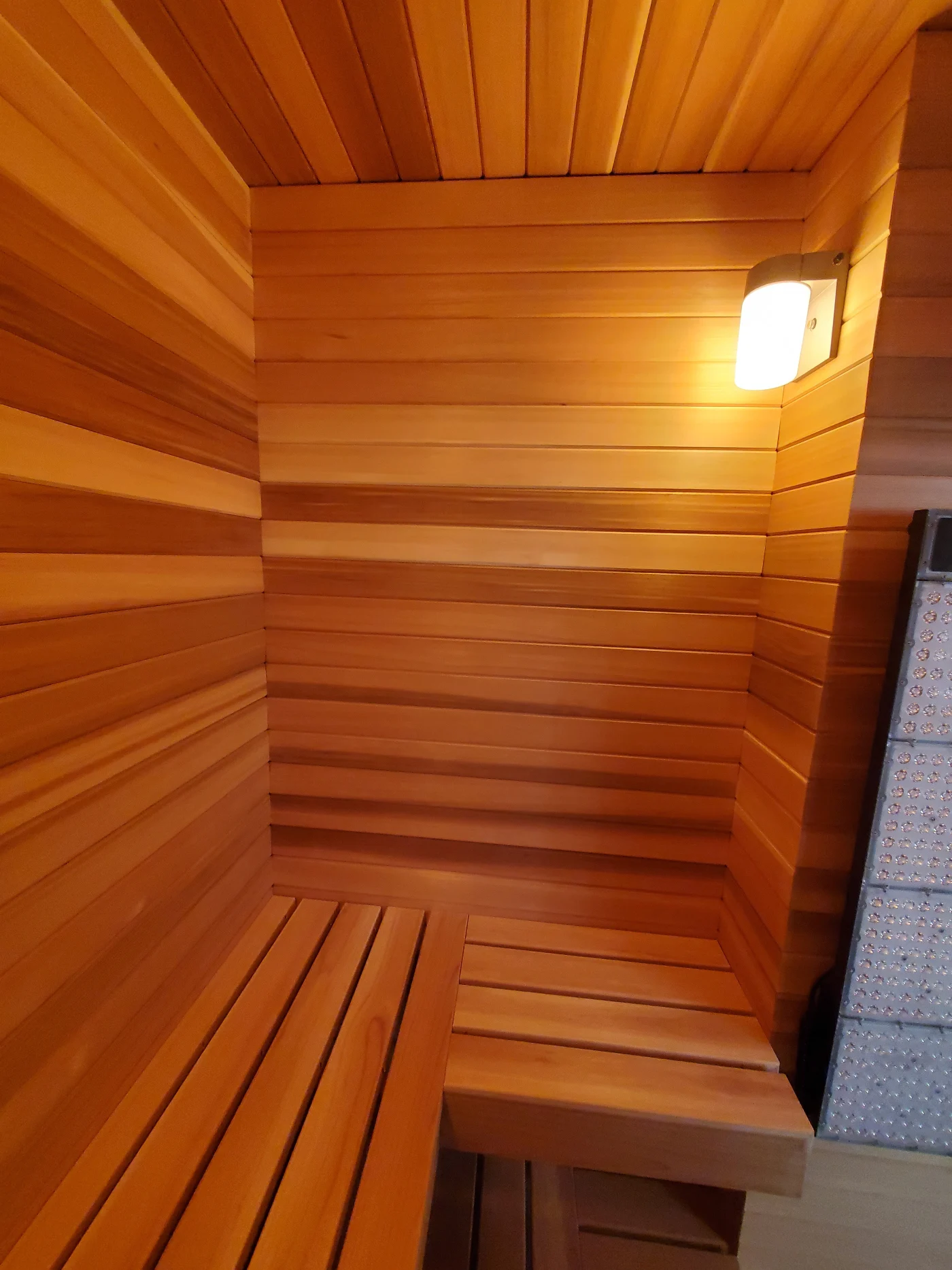 The interior of a sauna is covered in wooden paneling. The benches are made of the same color wood. There's a light mounted high on the right wall.