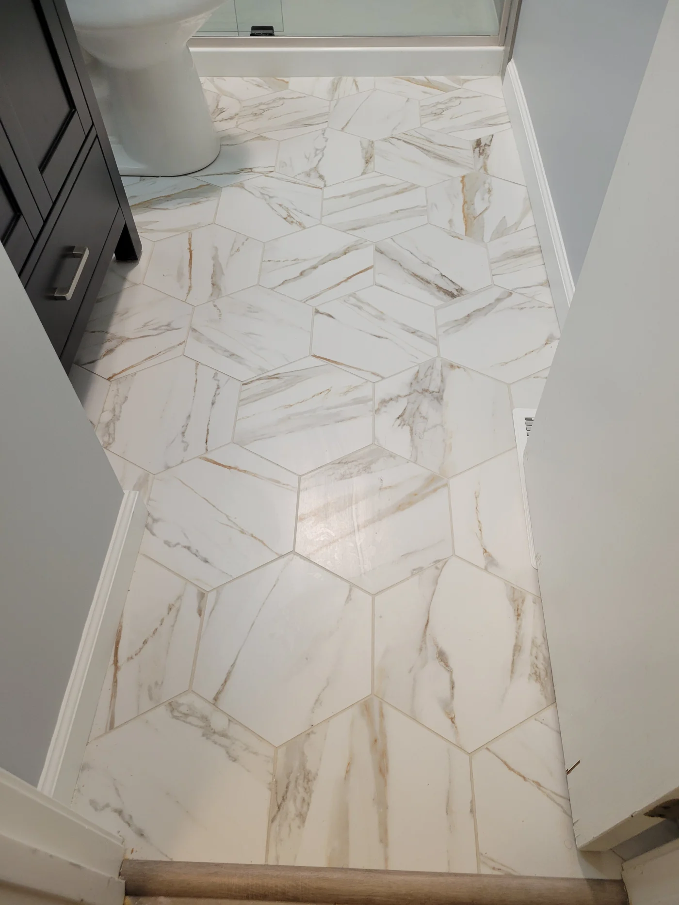 A remodeled bathroom floor with marble-look hexagon tiles beside a dark vanity, white toilet, glass shower, and wood threshold.