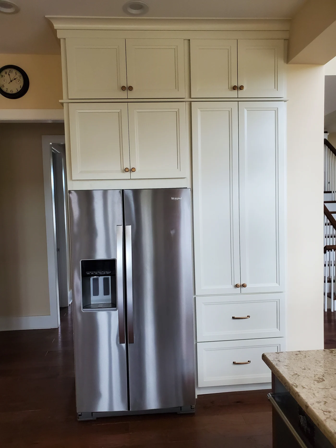 A remodeled kitchen with white floor-to-ceiling cabinets, stainless steel refrigerator, built-in pantry drawers, dark wood floors, and stone island.