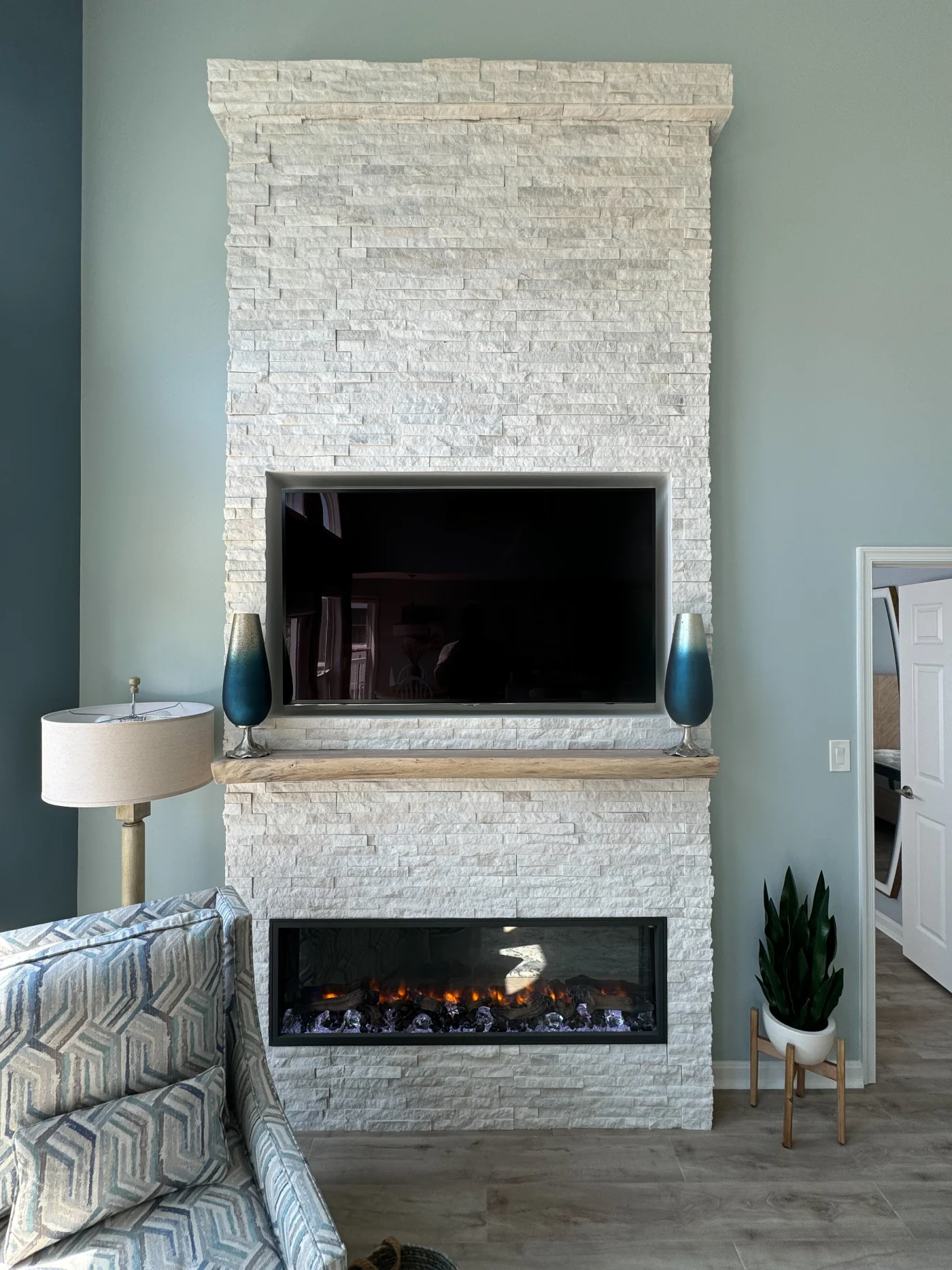 Remodeled living room with a white stacked-stone fireplace, mounted TV, wood mantel, electric firebox, blue walls, and gray flooring.