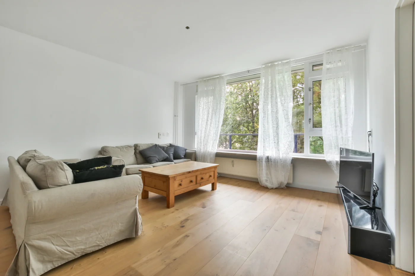 This bright living area features light hardwood flooring, large windows with sheer curtains, neutral seating, and clean white walls following an interior remodeling update.