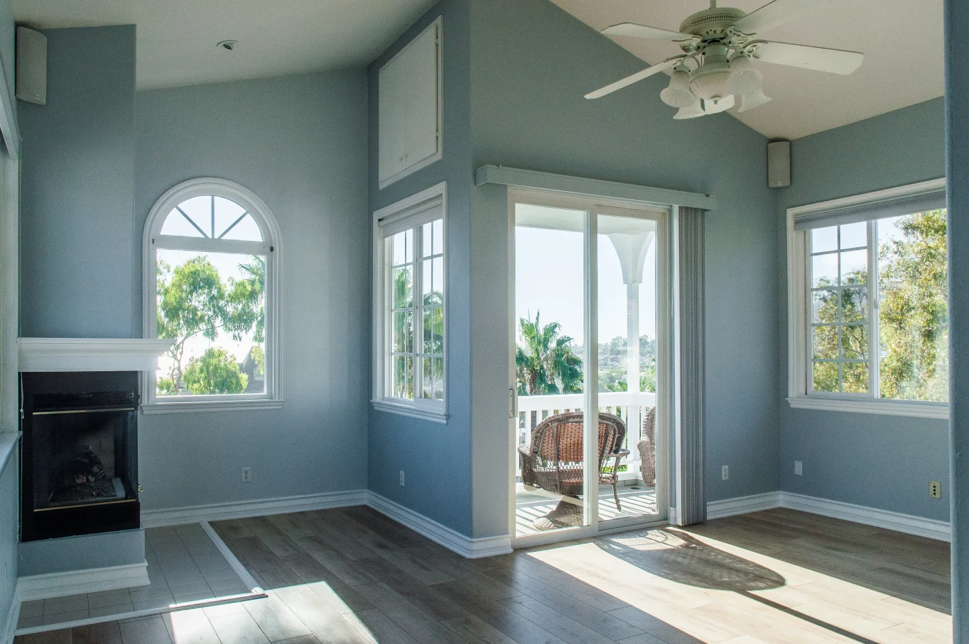 This sunlit living space features muted blue walls, a vaulted ceiling with a fan, a cozy fireplace, and sliding glass doors leading to a balcony, following a complete interior remodeling project.